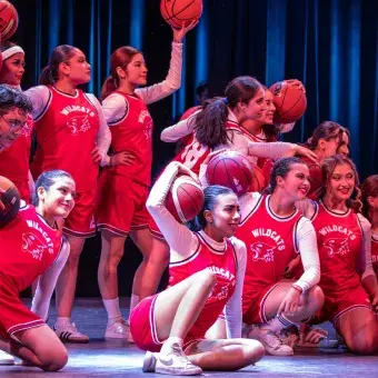 Estudiantes interpretan una coreografía con balones de baloncesto durante el número Get’cha Head in the Game del musical High School Musical: On Stage, con vestuario deportivo rojo.
