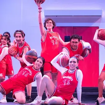 Estudiantes caracterizados como el equipo de baloncesto Wildcats posan con balones en escena durante High School Musical: On Stage, en una coreografía grupal del musical estudiantil.