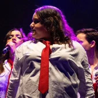 Chica vestida estilo colegial cantando en el escenario volteando hacia la izquerda.