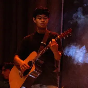 Chico tocando la guitarra acústica en escenario con humo alrededor. 
