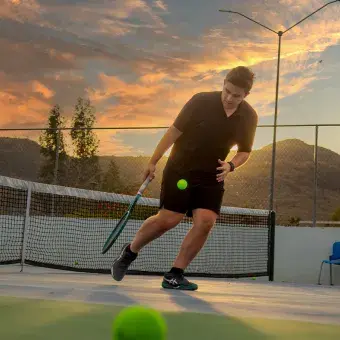 Joven universitario jugando tenis con atardecer de fondo.